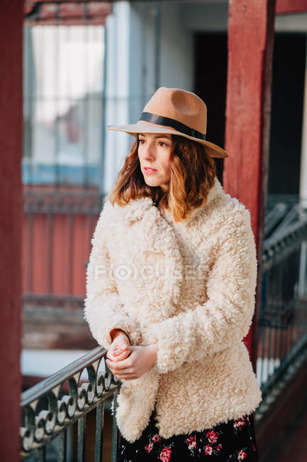 Positive attractive young woman in warm wear and hat looking away and standing near house and fence — Stock Photo