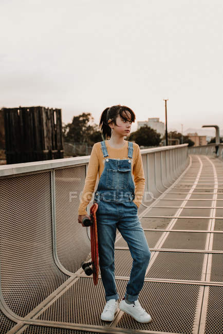Stylish teen girl in jean overall with hand in pocket holding skateboard on metal bridge in city — Stock Photo