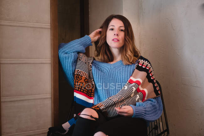 Belle jeune femme en pull tricoté avec écharpe regardant la caméra et assise sur une chaise près du mur dans la chambre — Photo de stock