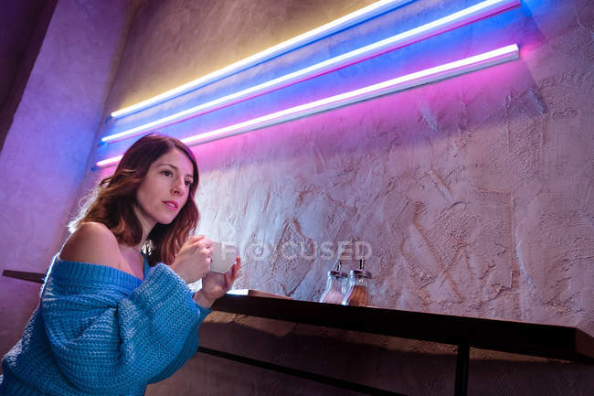 Удовлетворенная молодая женщина в трикотажном свитере держит чашку горячего напитка за столом у стены с неоновыми огнями — стоковое фото