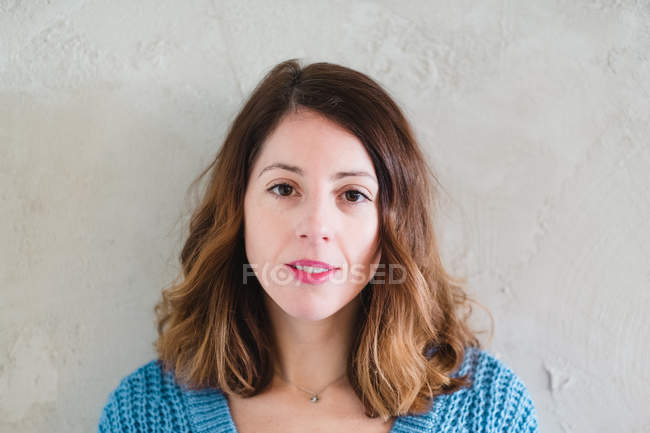 Belle jeune femme sérieuse en pull tricoté regardant la caméra et debout près du mur gris — Photo de stock