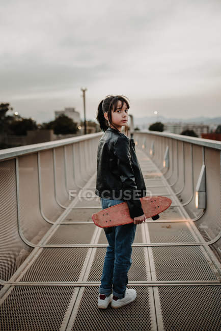 Adolescente chica con monopatín de pie en metal puente y mirando por encima del hombro - foto de stock