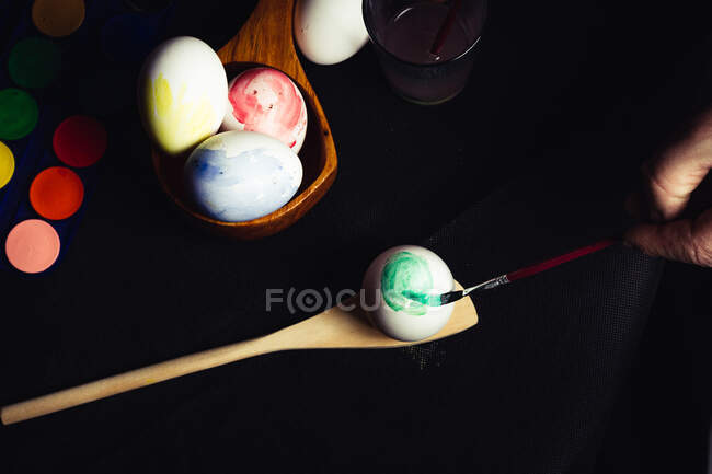 Сверху рука анонимного человека, использующего небольшую кисть для цвета хрупкого пасхального яйца на черном фоне — стоковое фото