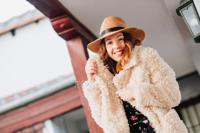 Позитивная привлекательная молодая женщина в теплой одежде и шляпе смотрит в камеру и стоит рядом с домом и забором — стоковое фото