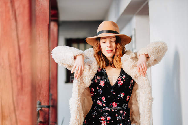 Позитивная привлекательная молодая женщина в теплой одежде и шляпе смотрит вдаль и стоит рядом с домом и забором — стоковое фото