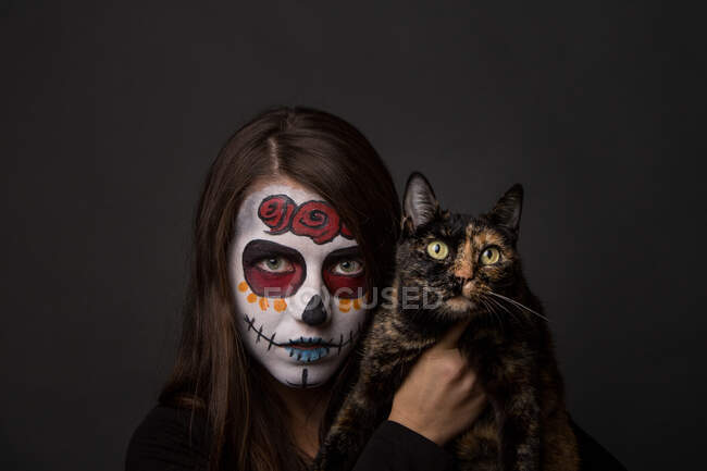 Jeune femme brune avec un maquillage sombre tenant chat drôle et regardant la caméra sur fond noir — Photo de stock