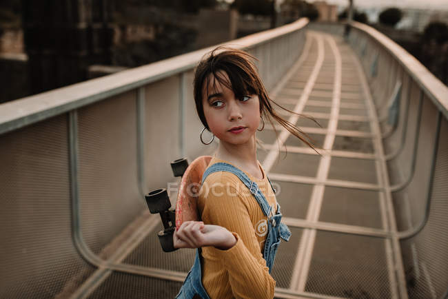 Chica con monopatín caminando sobre puente de metal y mirando por encima del hombro - foto de stock