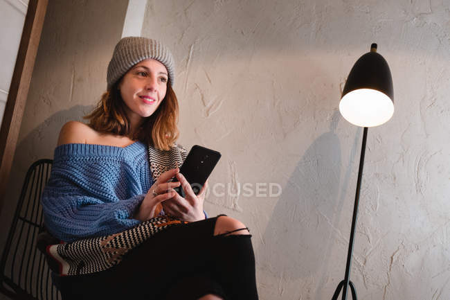 Молода жінка в трикотажному светрі з шарфом і капелюхом на мобільному телефоні і сидить на стільці біля стіни і лампи в кімнаті — стокове фото