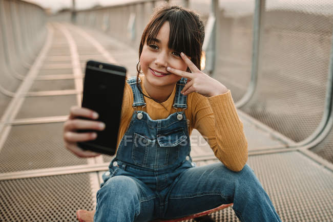 Fille drôle prenant selfie sur smartphone tout en étant assis sur planche à roulettes sur passerelle en métal — Photo de stock