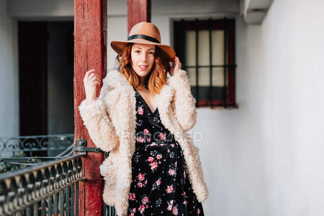 Mujer joven atractiva positiva en ropa abrigada y sombrero mirando hacia otro lado y de pie cerca de la casa y la cerca - foto de stock
