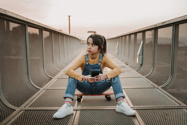 Дівчина зі смартфоном сидить на скейтборді на металевому мосту і дивиться вбік — стокове фото