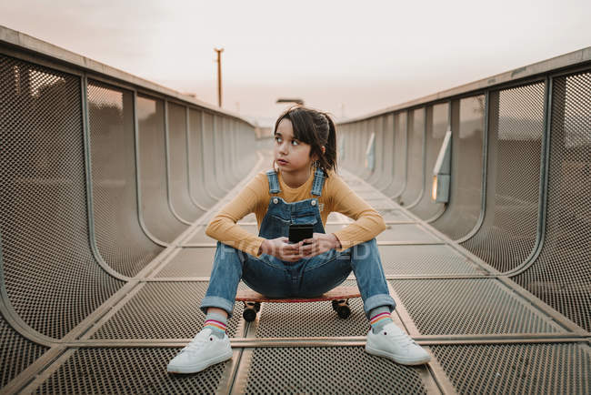 Chica con smartphone sentado en monopatín en puente de metal y mirando hacia otro lado - foto de stock