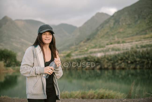 Жінка в спортивному одязі питна вода біля озера між горами — стокове фото