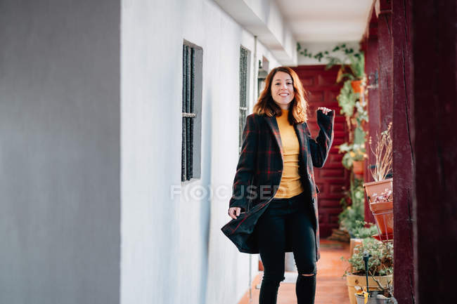 Mujer joven atractiva positiva mirando a la cámara y pasando por casa cerca de macetas con plantas - foto de stock