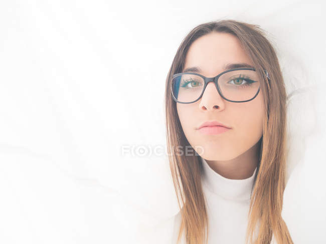 Orgoglioso adolescente in occhiali e usura bianca guardando la fotocamera tra le tende di luce — Foto stock