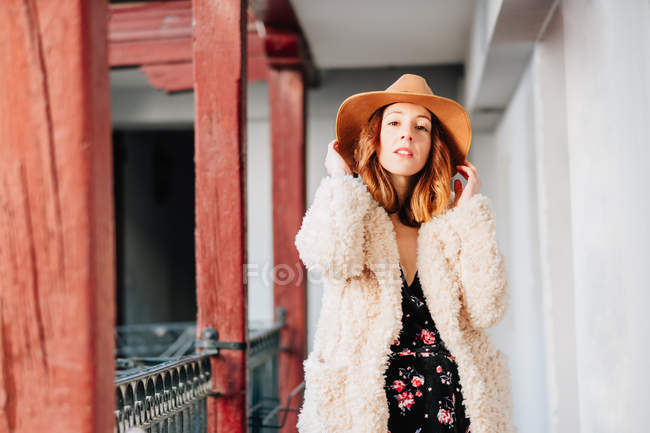 Positive attractive young woman in warm wear and hat looking at camera and standing near house and fence — Stock Photo