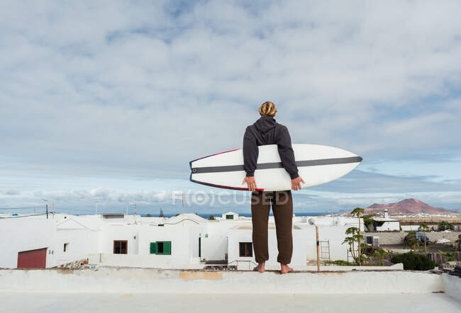Человек стоит с доской для серфинга на крыше здания и смотрит на горы — стоковое фото