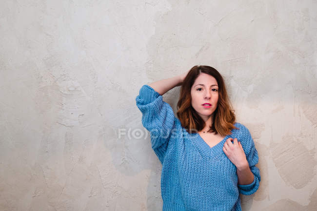 Милая серьезная молодая женщина в трикотажном свитере смотрит в камеру и стоит возле серой стены — стоковое фото