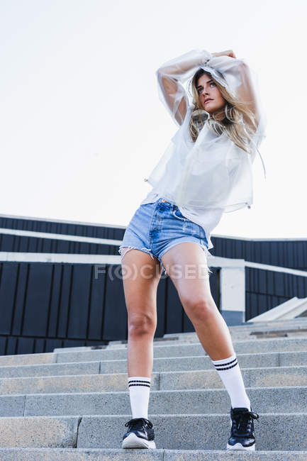 Sensual joven dama en impermeable, pantalones cortos de mezclilla y calcetines de rodilla posando en las escaleras en la calle - foto de stock