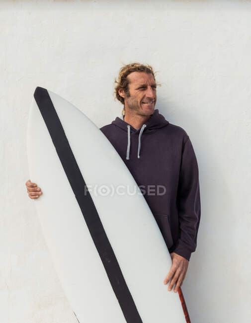 Bello adulto ragazzo in maglione in piedi con artigianato surf bordo vicino bianco muro — Foto stock