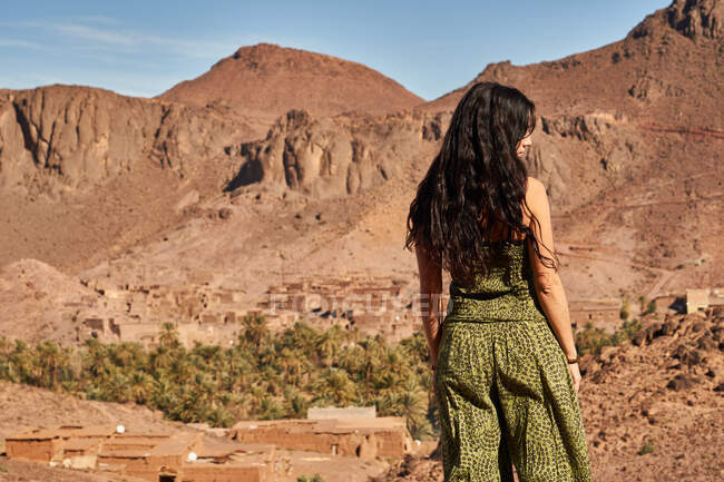 Vue arrière d'une jeune femme brune debout entre des terres désertiques près de constructions anciennes et de collines à Marrakech, au Maroc — Photo de stock