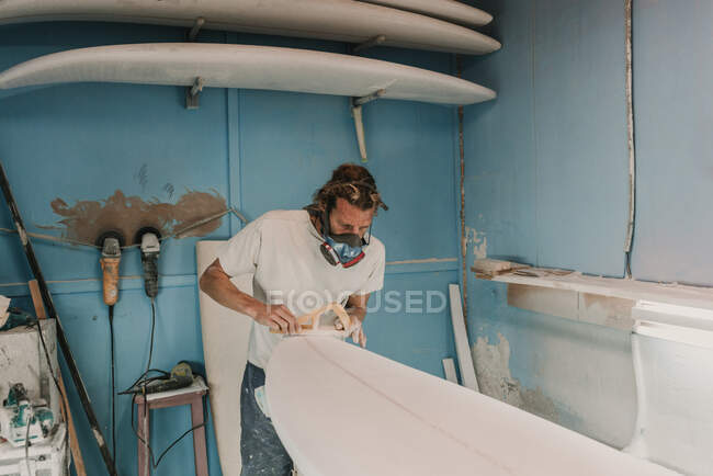 Mann mit Atemschutzmaske poliert Surfbrett in Werkstatt — Stockfoto