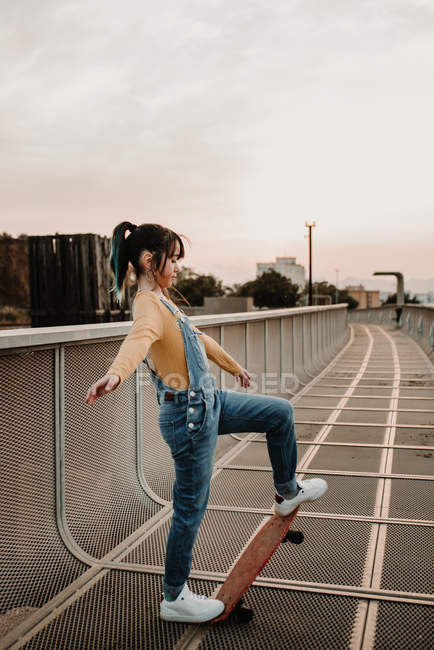 Chica de pie con monopatín en la pasarela de metal en la ciudad - foto de stock