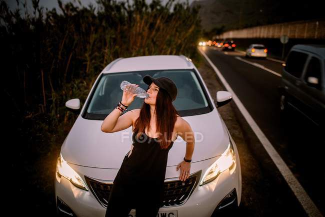 Mulher bebendo água de garrafa perto do carro na estrada à noite — Fotografia de Stock