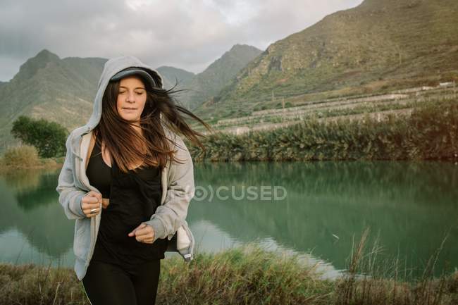 Mujer positiva en ropa deportiva corriendo en la naturaleza cerca del lago y las montañas - foto de stock