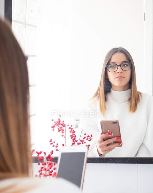 Reflection of proud teen in eyeglasses and white wear holding mobile phone near mirror — Stock Photo