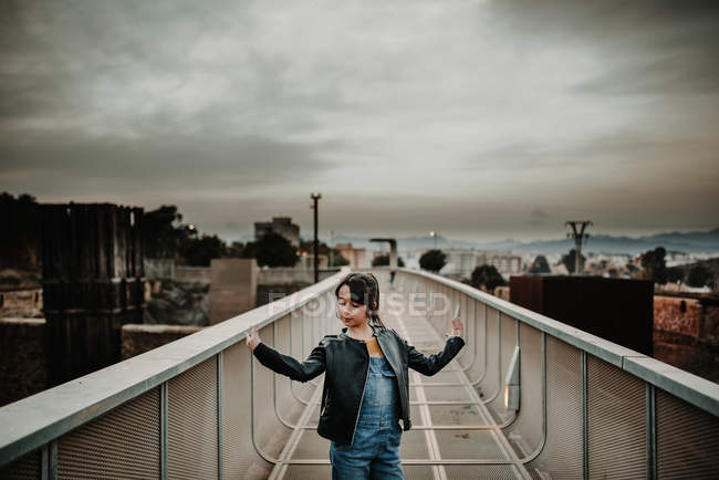 Stylish teen girl in jean overall and leather jacket posing on metal bridge in city under cloudy sky — Stock Photo