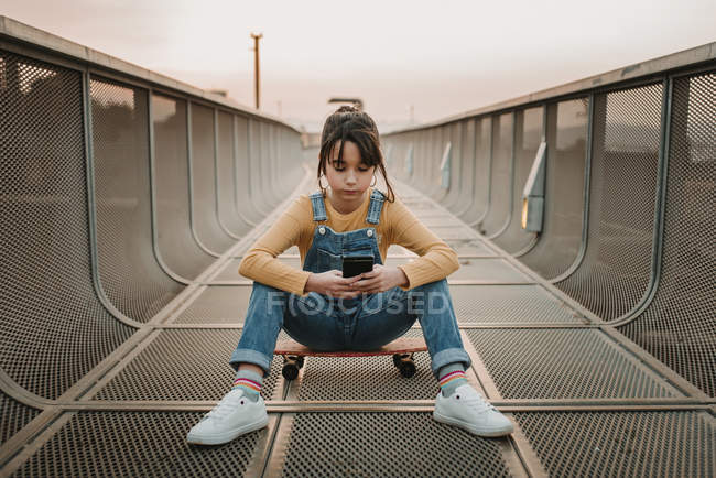 Stylish teen girl in jean overall sitting on skateboard and using mobile phone on metal bridge in city — Stock Photo