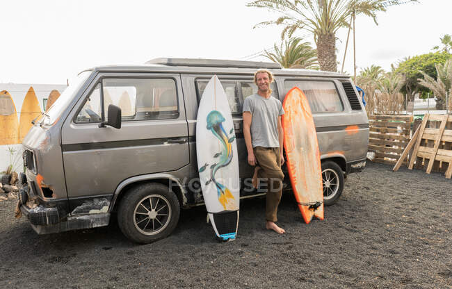 Hombre adulto feliz de pie entre tablas de surf artesanales y camioneta vieja en el patio - foto de stock