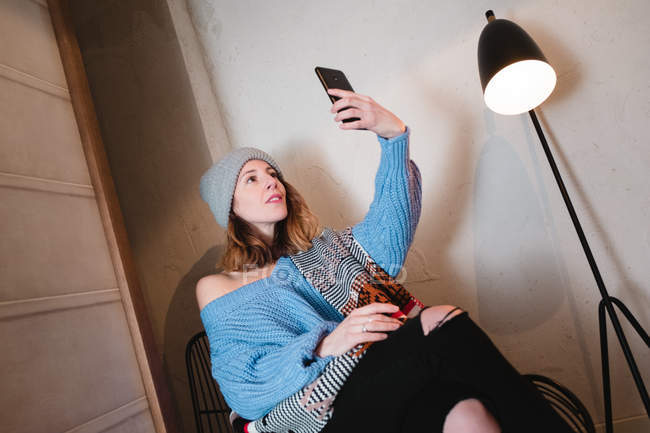 Young woman in knitted sweater with scarf and hat taking selfie on smartphone and sitting on chair near wall and lamp in room — Stock Photo