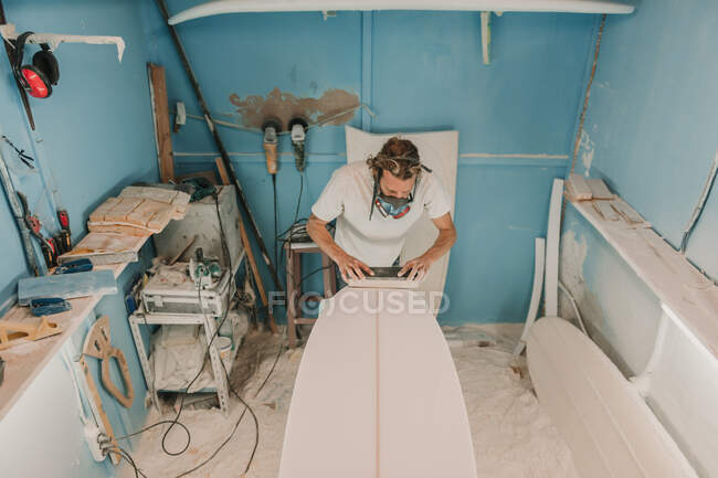Man in respirator polishing surf board in workshop — Stock Photo