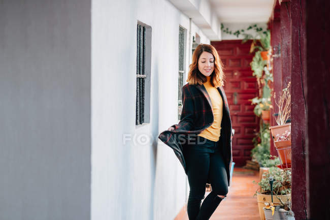 Positif attrayant jeune femme va sur le passage dans la maison près de pots de fleurs avec des plantes — Photo de stock