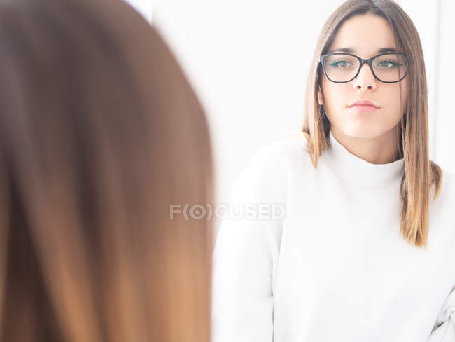 Reflection of proud teen in eyeglasses and white wear looking at camera near window — Stock Photo