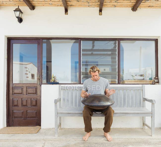 Homme pieds nus en t-shirt assis sur le siège avec grand tambour à main près de la porte et les fenêtres du bâtiment — Photo de stock