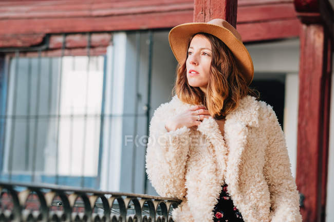 Positive attractive young woman in warm wear and hat looking away and standing near house and fence — Stock Photo
