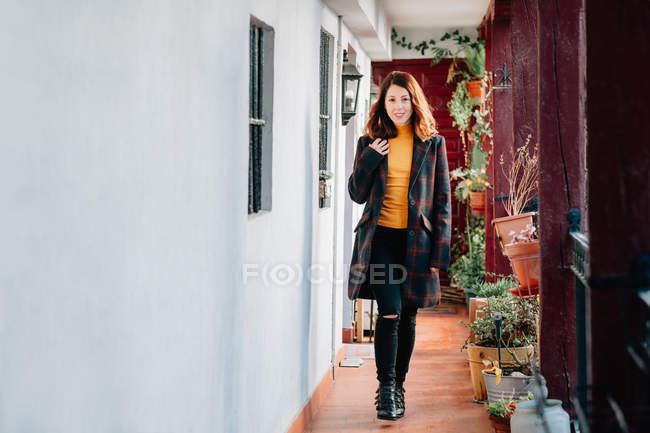 Позитивная привлекательная молодая женщина в теплой одежде, смотрящая в камеру и проходящая по проходу в доме рядом с цветочными горшками с растениями — стоковое фото