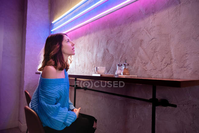 Позитивная женщина с чашкой за столом у стены с неоновыми лампами — стоковое фото