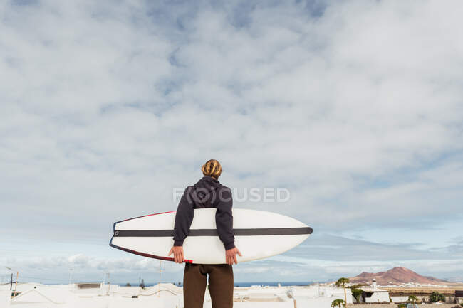 Hombre de pie con tabla de surf en el techo del edificio y mirando a la montaña - foto de stock