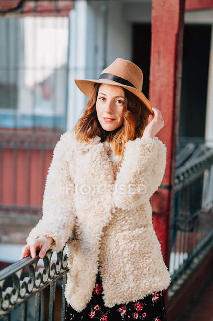 Позитивна приваблива молода жінка в теплому одязі і капелюсі дивиться на камеру і стоїть біля будинку і паркану — стокове фото