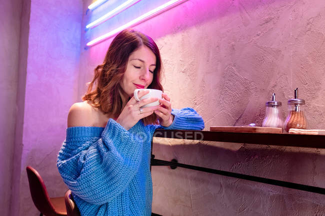 Pleased young woman in knitted sweater with closed eyes holding cup of hot drink at table near wall with neon lights — Stock Photo