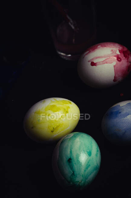 Зверху набір погано пофарбованих яєць різних кольорів, розташованих біля склянки води на чорному тлі — стокове фото