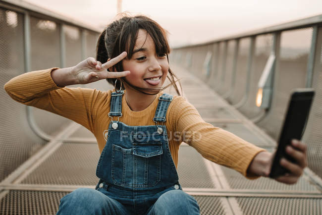 Chica alegre tomando selfie con teléfono inteligente en la pasarela de metal - foto de stock