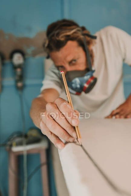 Mann mit Atemschutzmaske misst Surfbrett in Werkstatt — Stockfoto
