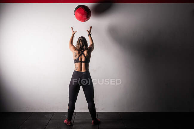 Atlética joven dama concentrada en ropa deportiva en tiza levantando pesada pelota de medicina en el hombro en el gimnasio - foto de stock