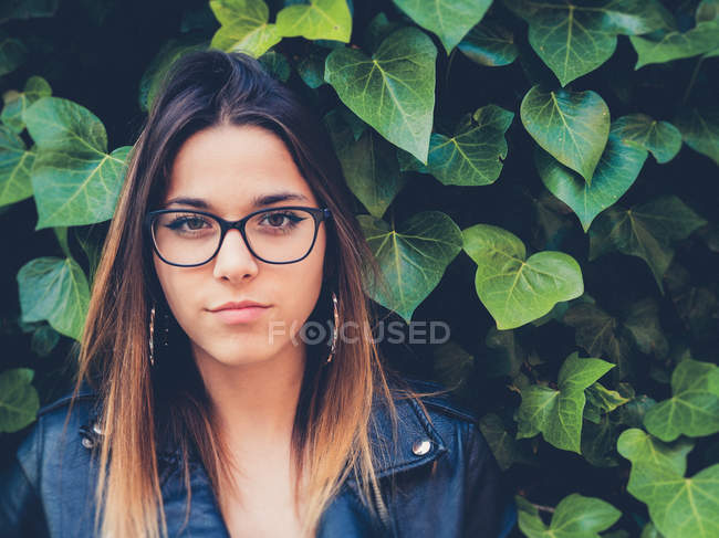 Adolescente in occhiali e giacca di pelle guardando la fotocamera vicino al fogliame verde di arbusto — Foto stock