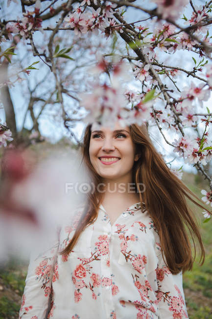 Vista attraverso ramoscelli di albero da frutto in fiore di attraente signora allegra guardando lontano in giardino — Foto stock